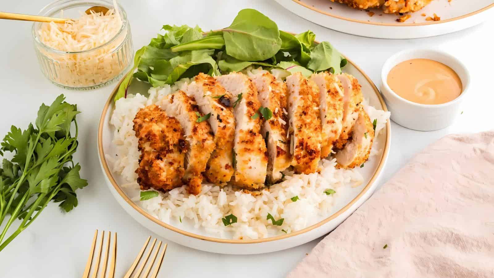 A plate of sliced breaded chicken on white rice with greens, accompanied by a small bowl of sauce and a jar of grated cheese.