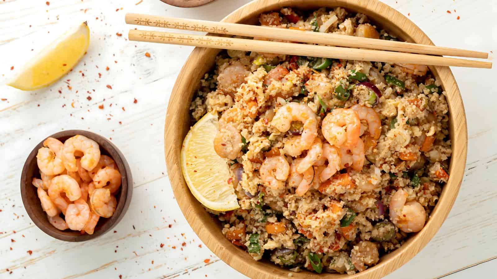 A bowl of couscous with shrimp, vegetables, and a lemon wedge, accompanied by chopsticks, is next to a small dish of shrimp on a white wooden surface.