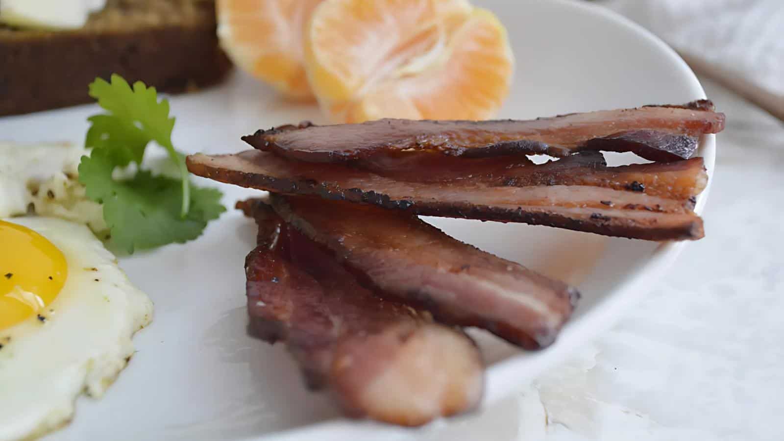 Close-up of a breakfast plate with three strips of cooked bacon, a sunny-side-up egg, a sprig of cilantro, and orange slices on a white plate.