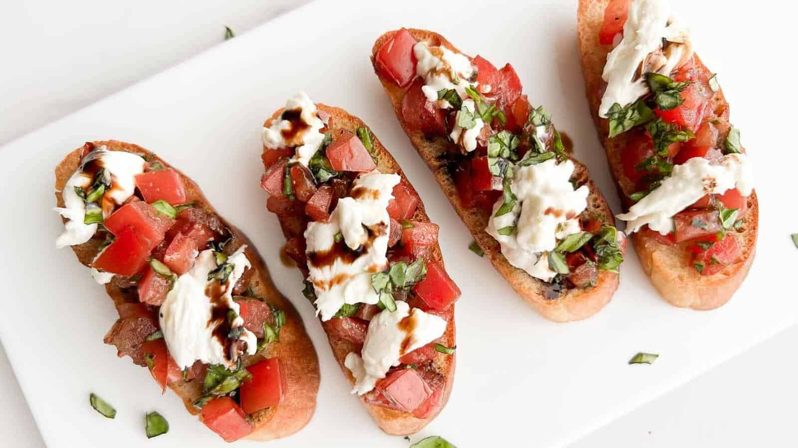 Four bruschetta slices topped with chopped tomatoes, mozzarella, basil, and balsamic glaze on a white rectangular plate.