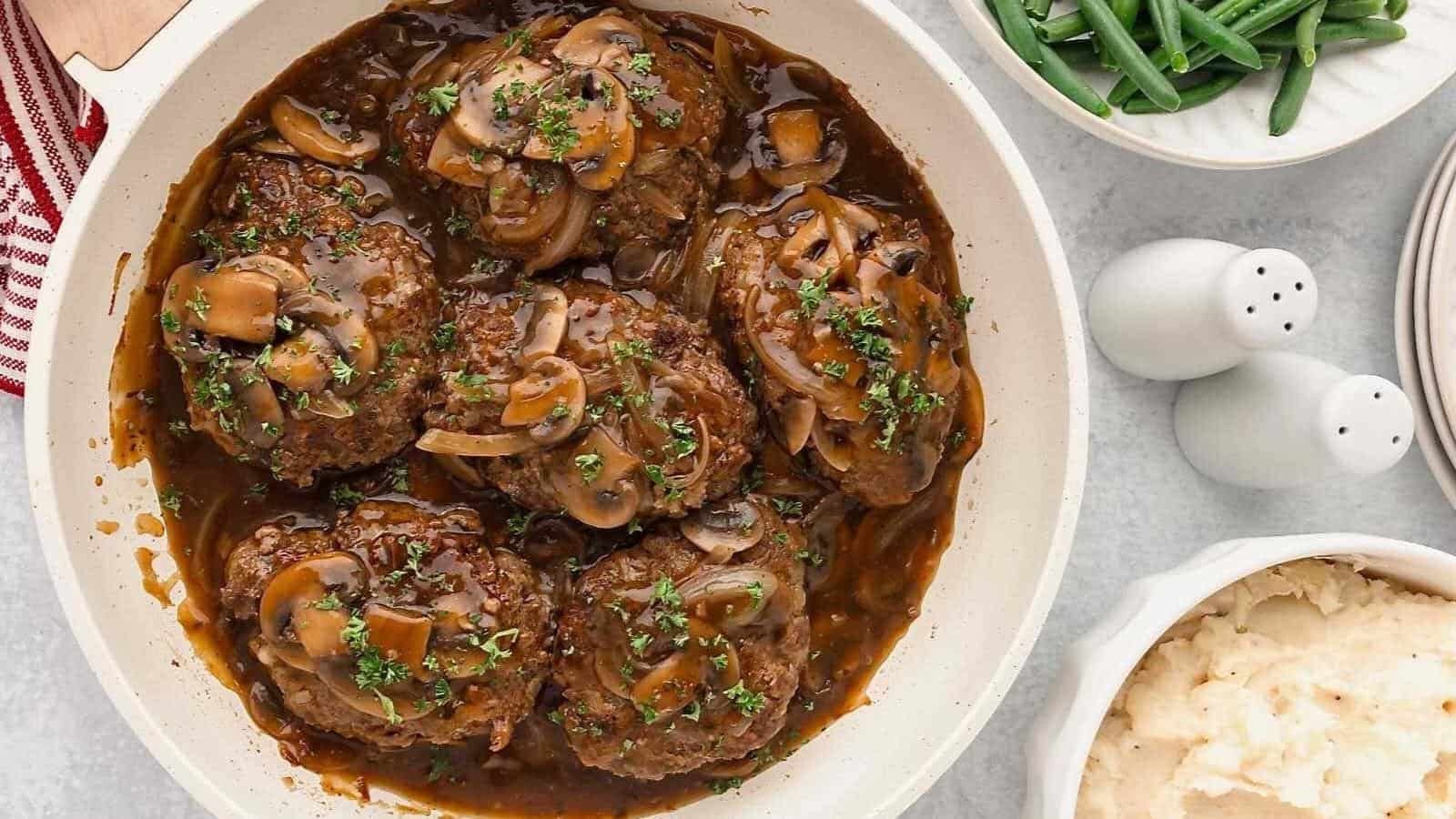 A dish of Salisbury steak with mushroom gravy, garnished with parsley, next to bowls of green beans and mashed potatoes, with salt and pepper shakers nearby.