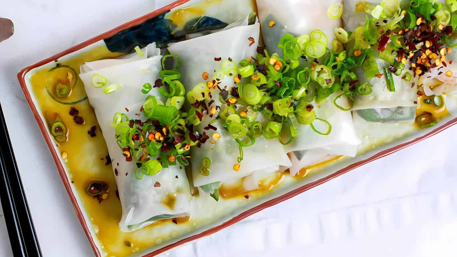 A tray of steamed rice paper rolls topped with scallions, chili flakes, and a sauce, served on a rectangular dish.