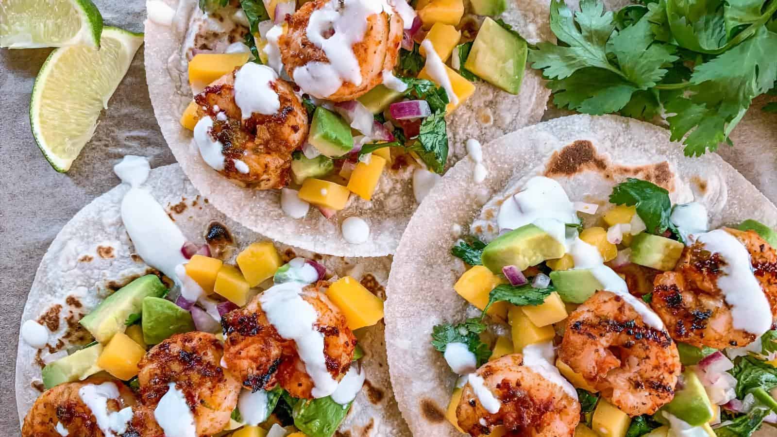 Three shrimp tacos topped with avocado, diced mango, cilantro, onions, and drizzled with a white sauce. Lime wedges are on the side.