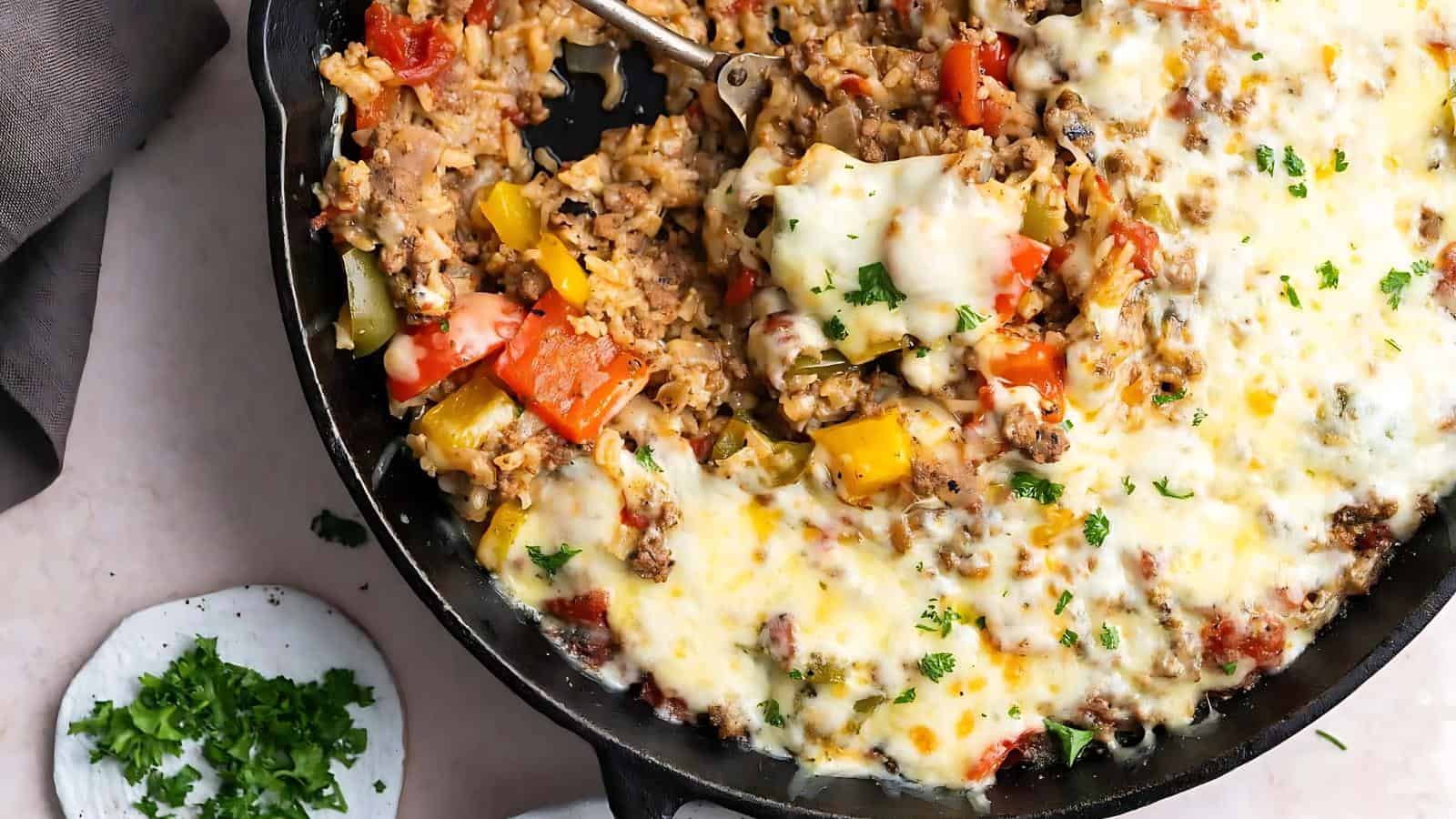 A skillet filled with a rice and vegetable casserole topped with melted cheese. A portion is removed, showing a serving spoon. A small plate with chopped herbs is beside the skillet.