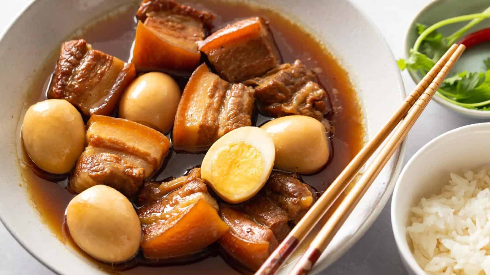 A bowl of braised pork belly and boiled eggs in soy sauce, served next to white rice and garnished with herbs and a red chili. Chopsticks rest on the plate's edge.