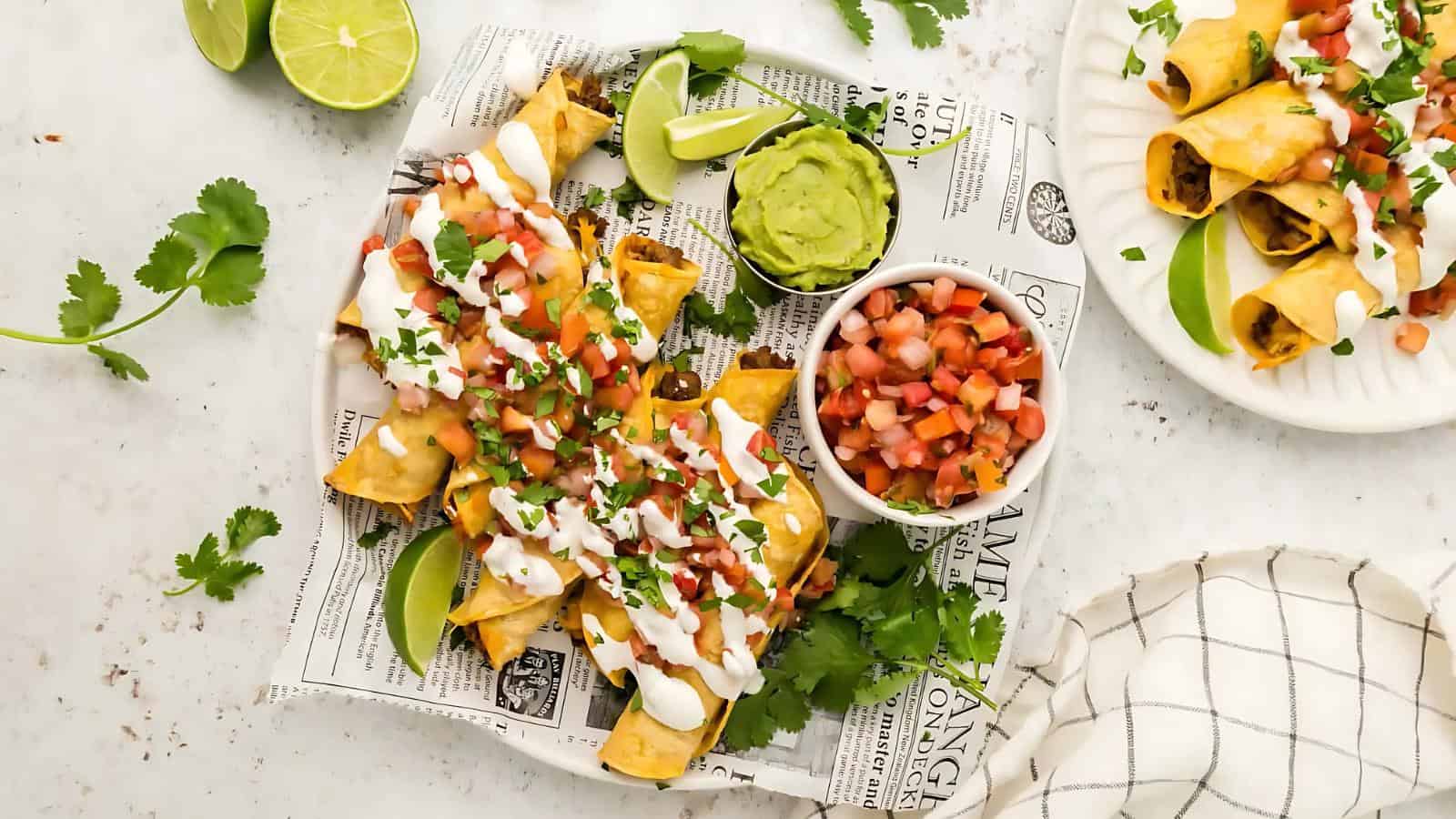 Plate of taquitos with sour cream, diced tomatoes, and cilantro, served with guacamole, salsa, and lime wedges on a newspaper-lined tray.