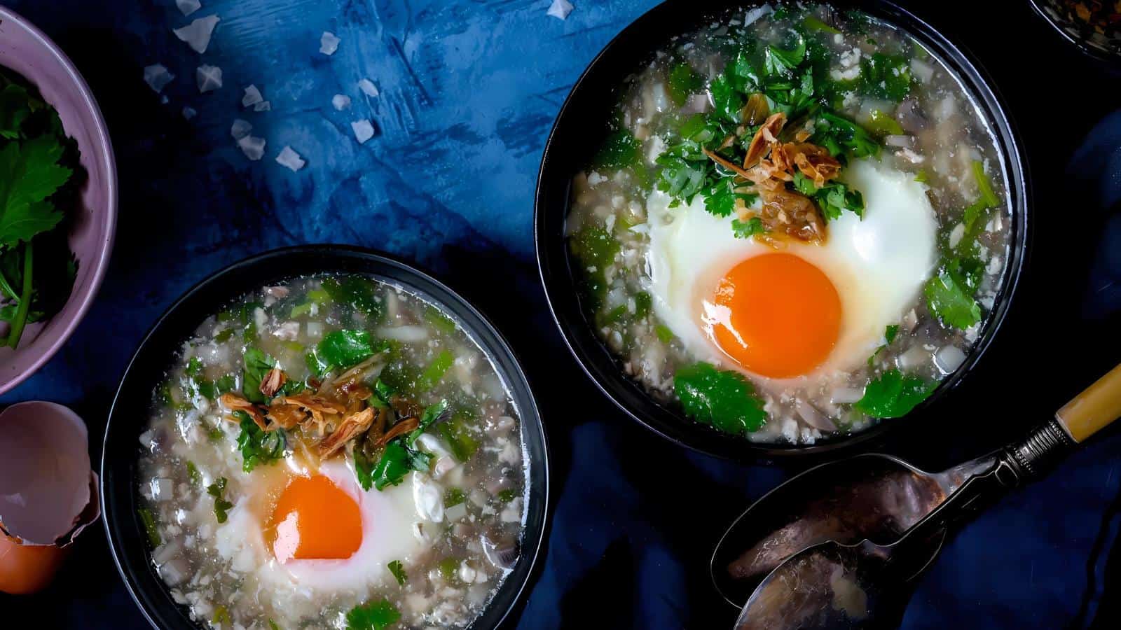 Two bowls of soup with poached eggs, greens, and fried onions on a dark blue surface. An empty shell and spoons are nearby.