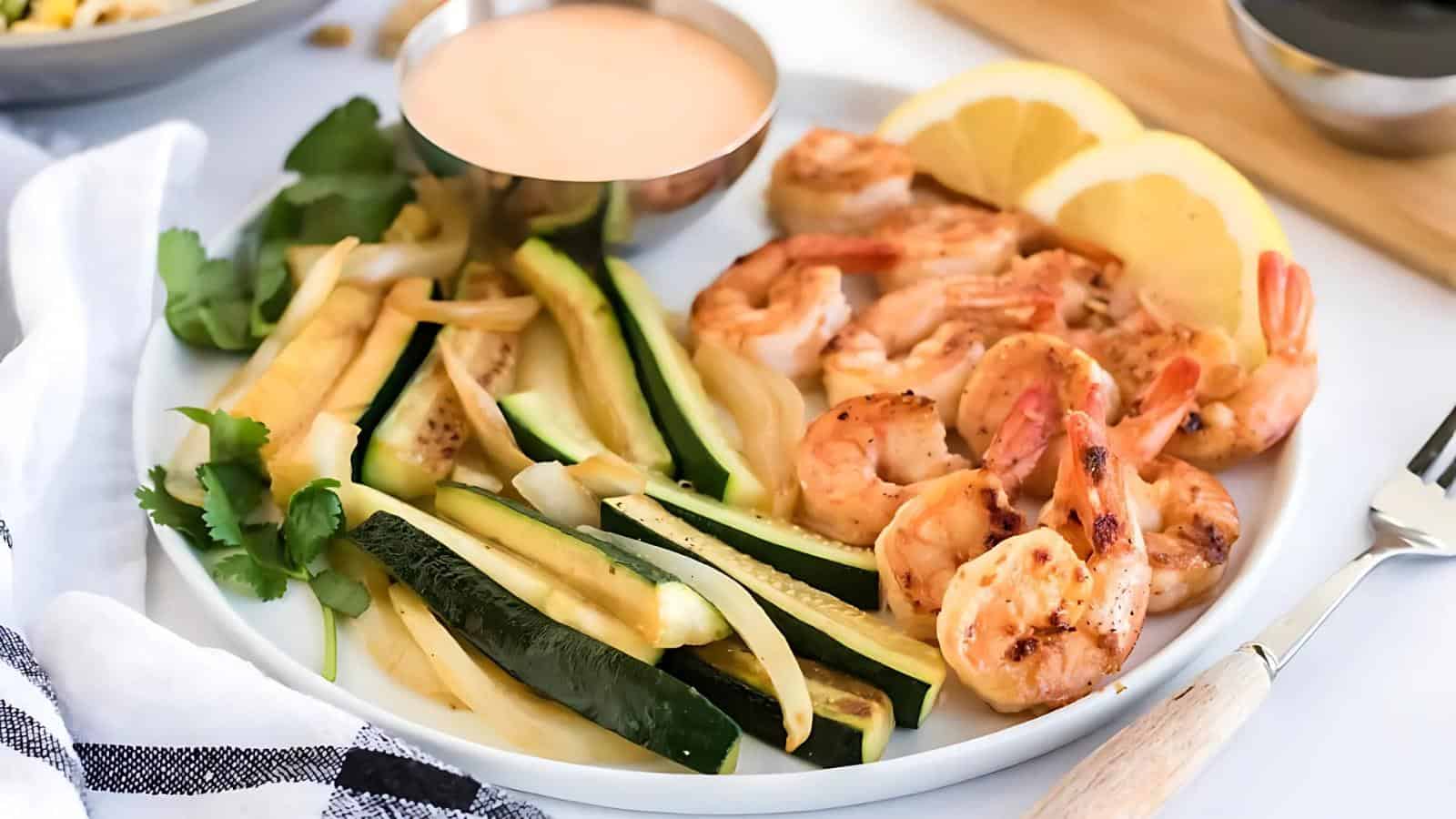 Plate with grilled shrimp, sliced zucchini, lemon wedges, and a small bowl of sauce.