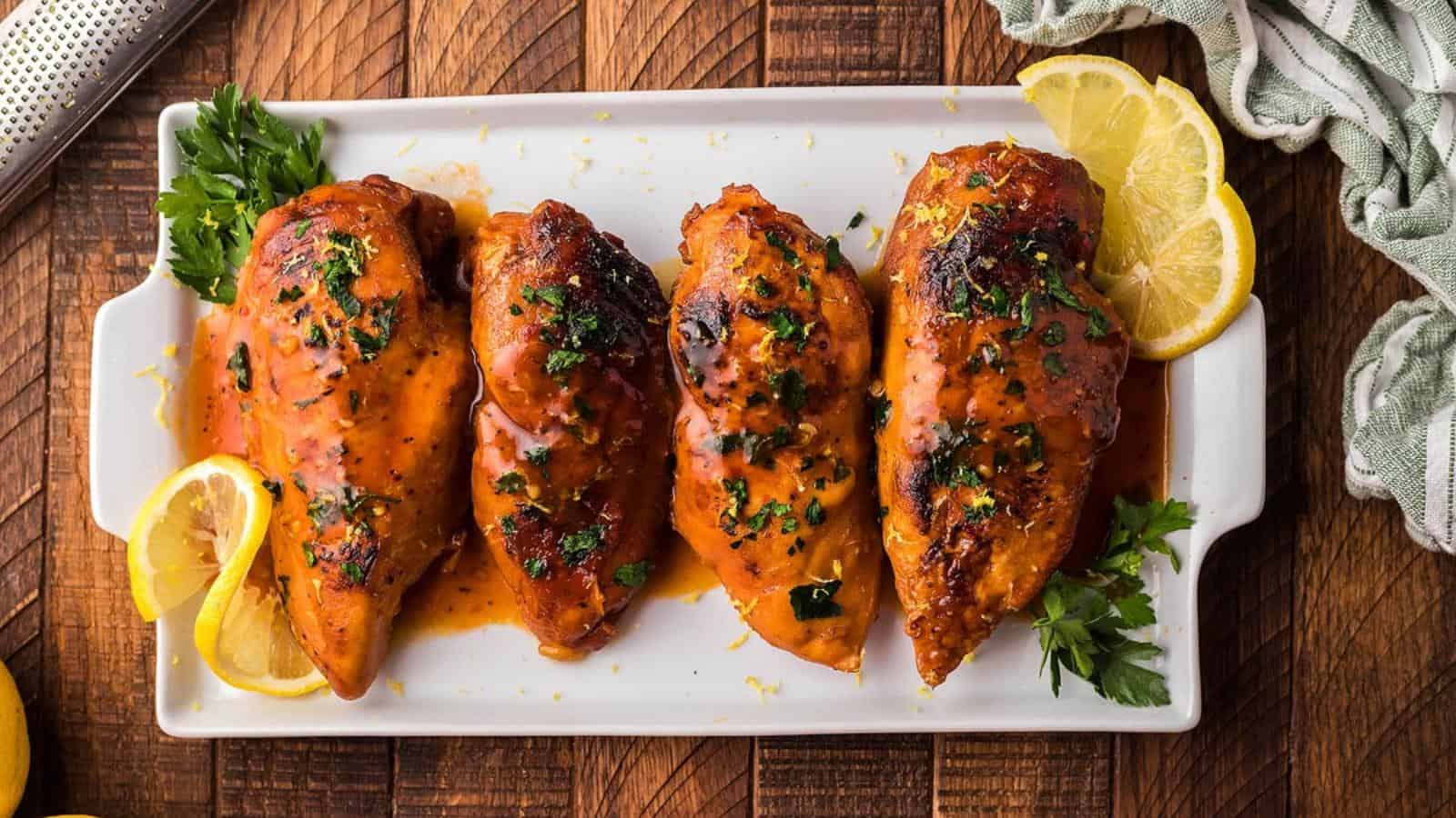 Four seasoned and roasted chicken breasts on a white platter, garnished with fresh herbs and lemon slices.