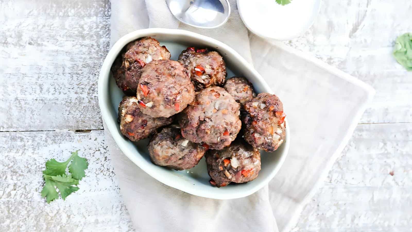 A white dish filled with cooked meatballs, garnished with red and white bits, placed on a light-colored table with a beige napkin and a small bowl of white sauce nearby.