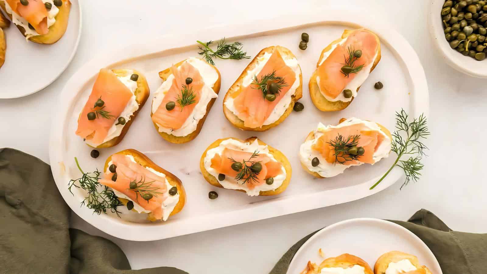 Plate of smoked salmon canapés with cream cheese, capers, and dill arranged on a white platter. Green napkin and a side dish of capers accompany the display.