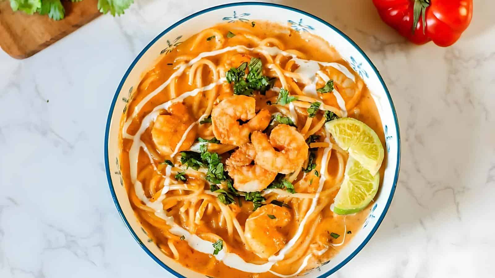 A bowl of creamy shrimp soup garnished with herbs and lime wedges, served with noodles.