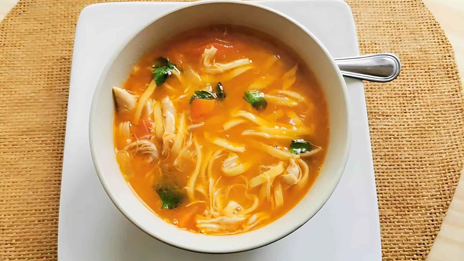 A bowl of chicken soup with shredded chicken, vegetables, and herbs on a white square plate and woven mat.