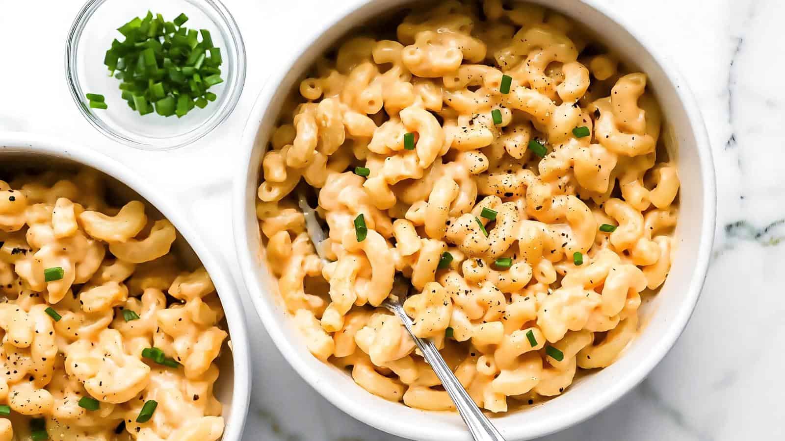Bowl of creamy macaroni and cheese topped with chopped chives, with a fork resting in it. A small bowl of chives is on the side.