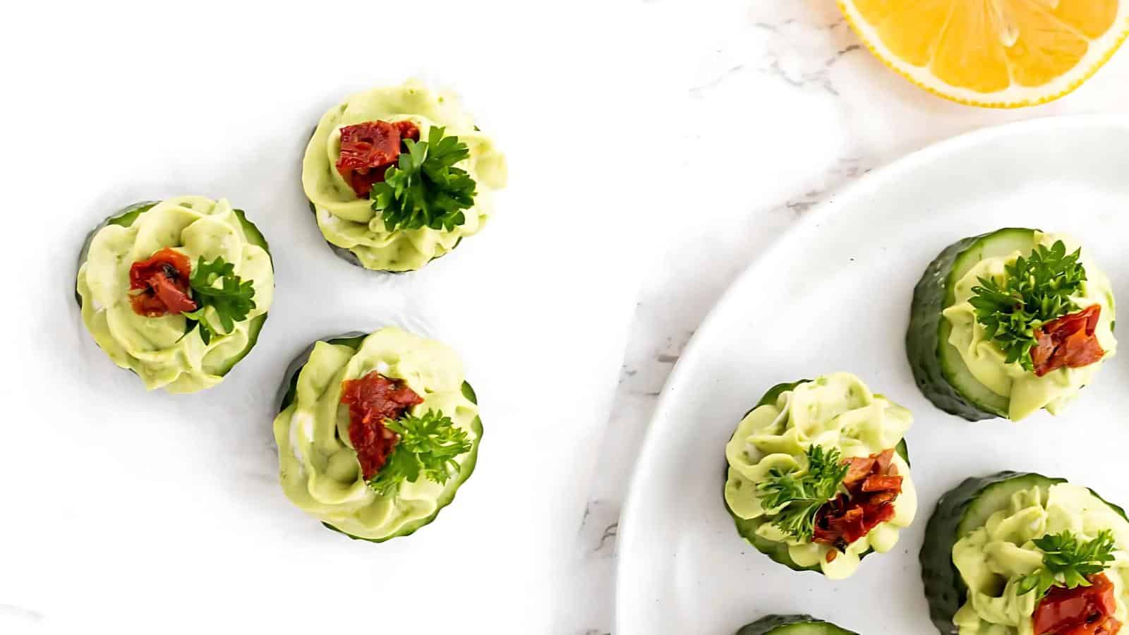 Cucumber slices topped with avocado spread, parsley, and sun-dried tomatoes on a plate with a lemon slice nearby.