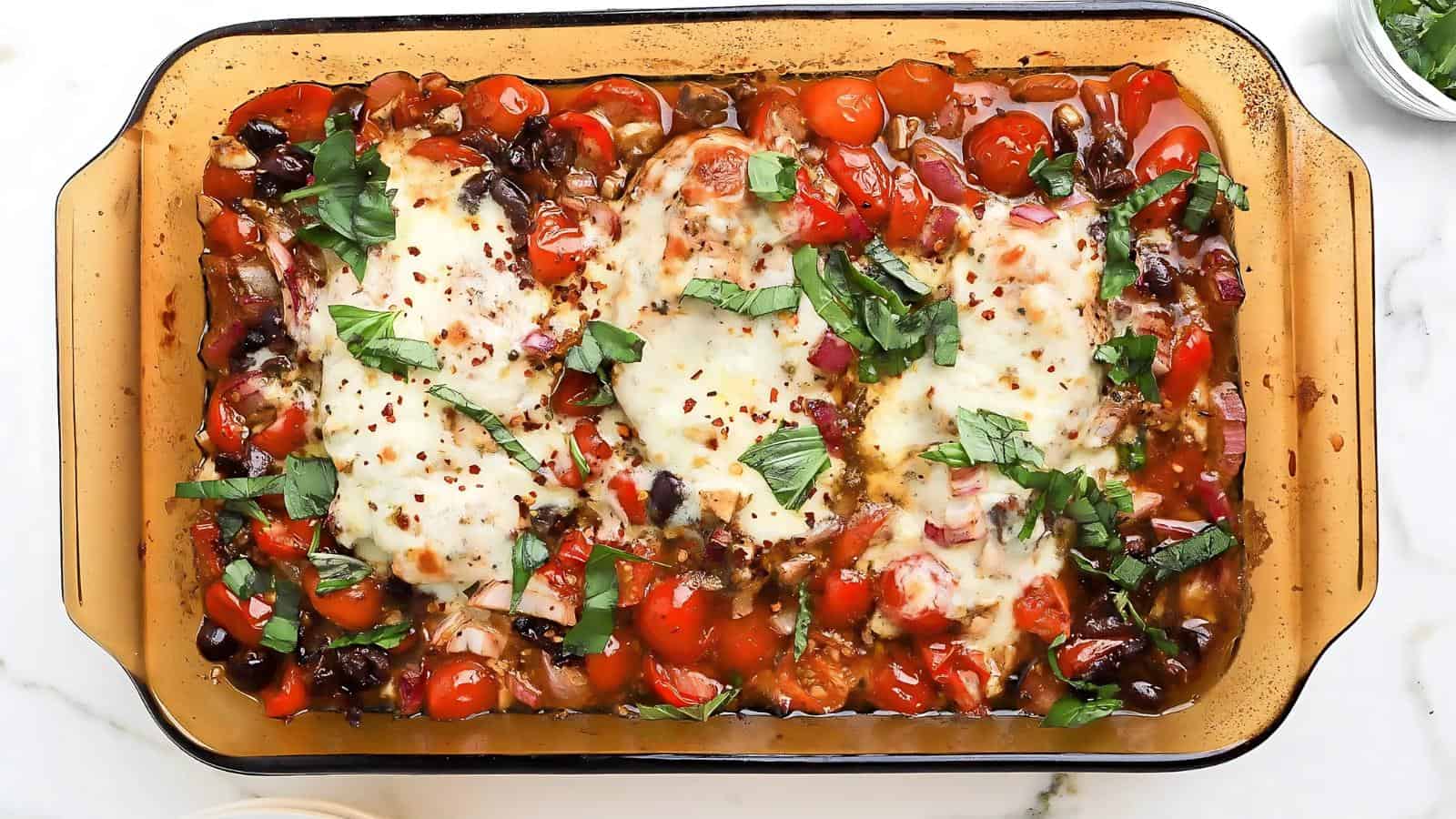 Baked dish with melted cheese on chicken breasts, surrounded by tomatoes, olives, and basil in a glass baking dish.