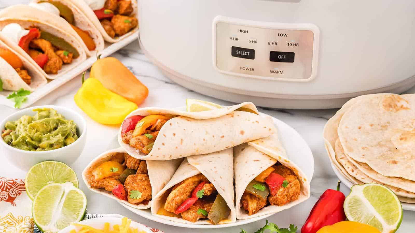 Three chicken fajitas with bell peppers on a plate near a slow cooker, surrounded by lime slices, guacamole, and tortillas.