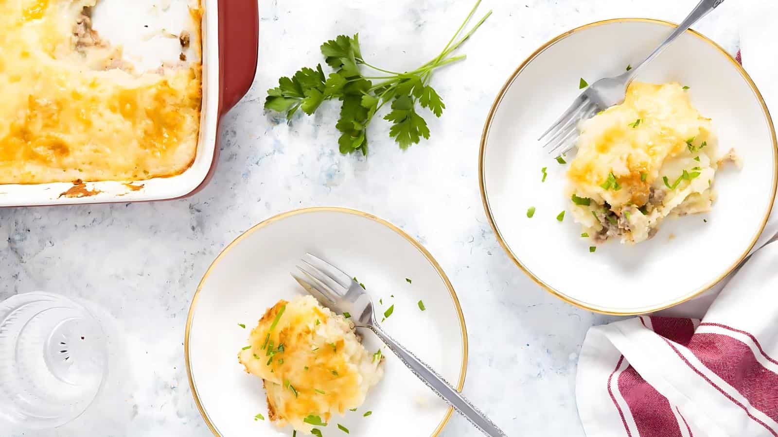 Two plates with servings of shepherd's pie garnished with parsley, a baking dish with more pie, a sprig of parsley, and a red-striped napkin on a white surface.