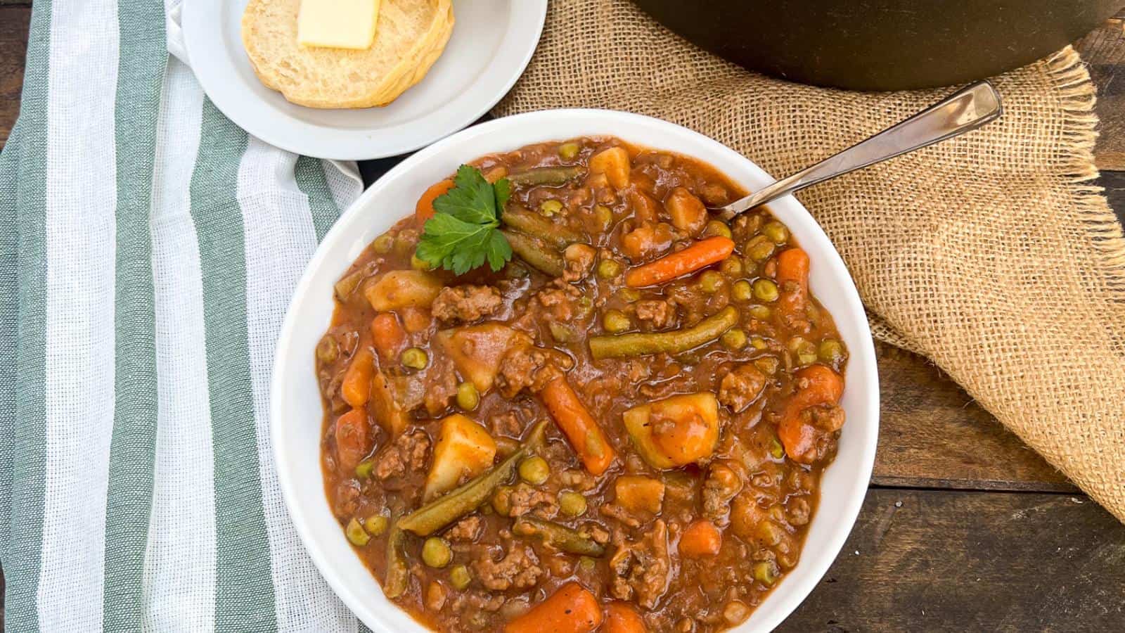 A bowl of stew with beef, potatoes, carrots, peas, and green beans. A plate with a buttered biscuit is beside the bowl, on a wooden table with a striped cloth and burlap.