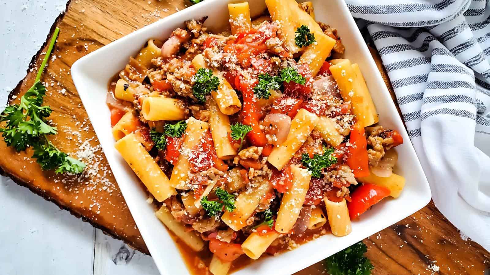 Square dish with pasta, chunks of vegetables, and parsley garnish. Sprinkled with grated cheese. Striped cloth and parsley on a wooden surface.