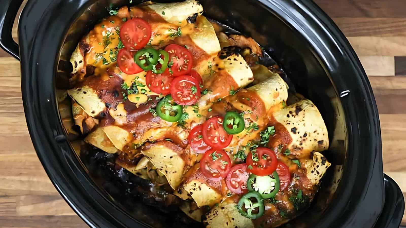 Enchiladas in a slow cooker, topped with melted cheese, sliced cherry tomatoes, jalapeños, and chopped herbs.