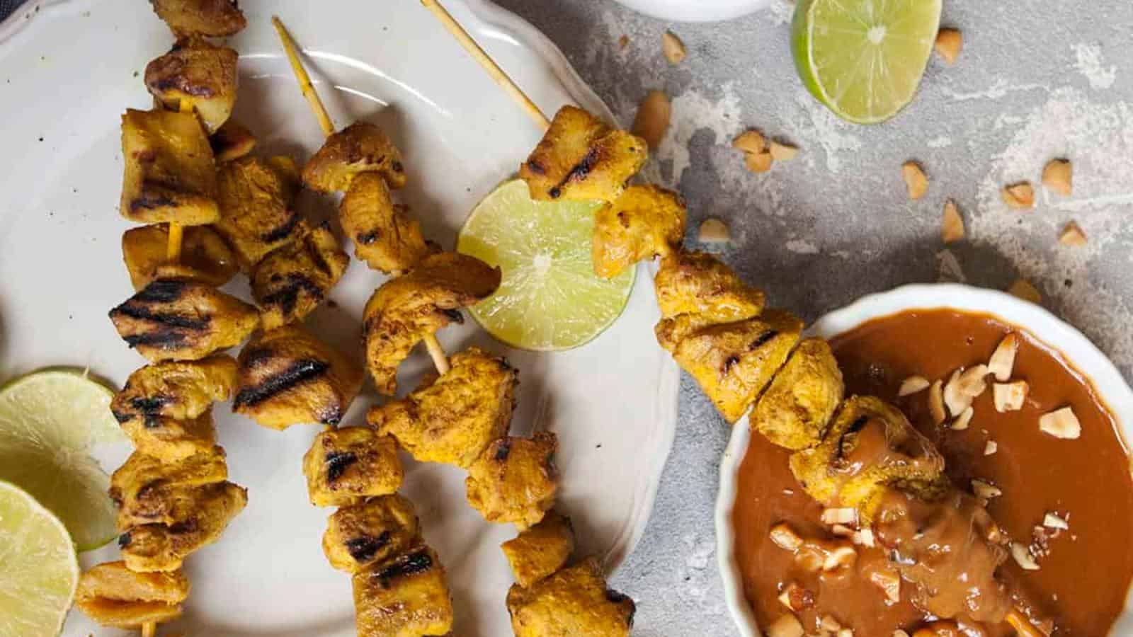 Grilled chicken skewers on a plate with lime slices beside a bowl of peanut sauce topped with chopped peanuts.