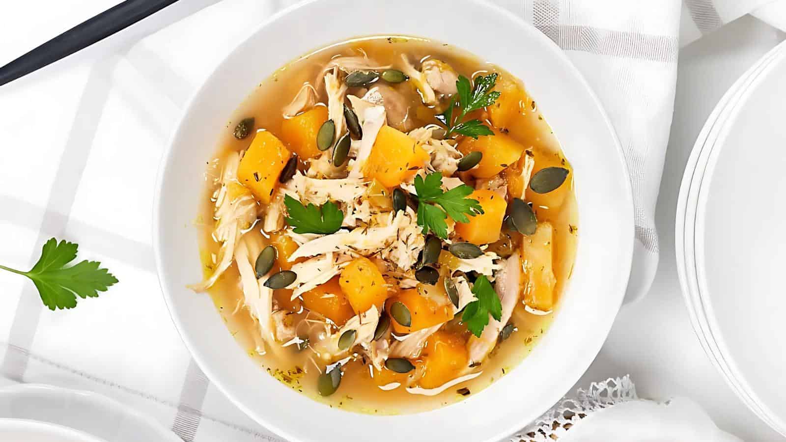 A bowl of soup with shredded chicken, diced squash, pumpkin seeds, and parsley garnish.
