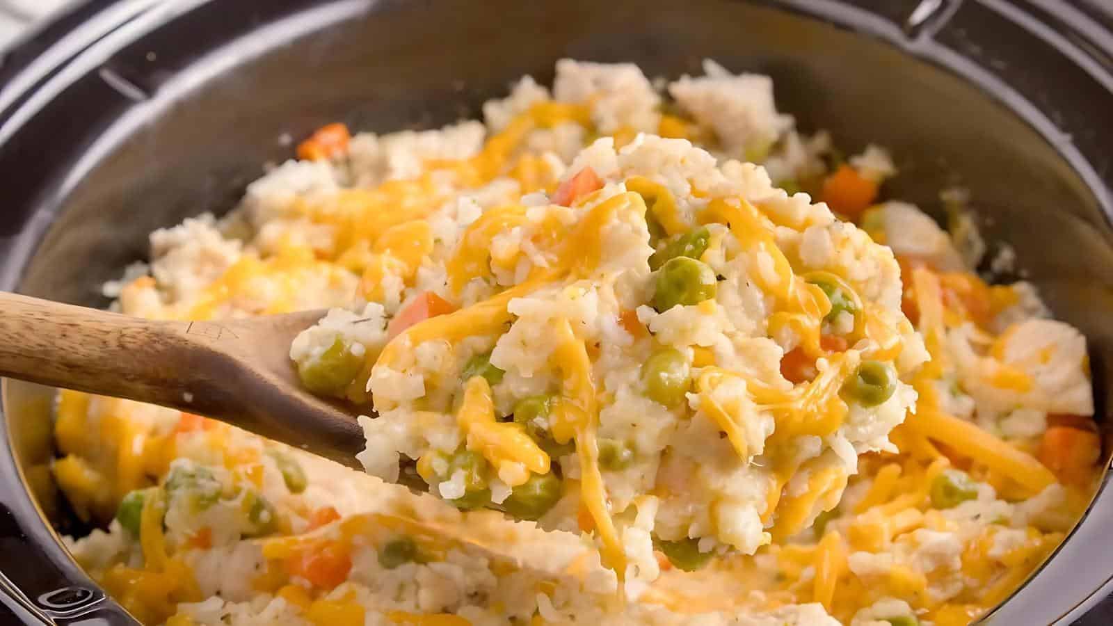 A wooden spoon scoops chicken, rice, peas, and carrots topped with melted cheese from a black cooking pot.