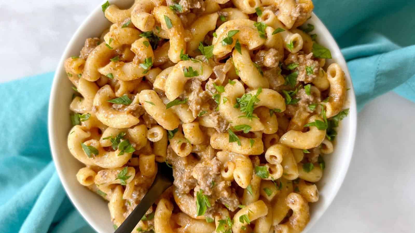 A bowl of macaroni mixed with ground meat and garnished with chopped herbs. A black fork is on the left side of the bowl.