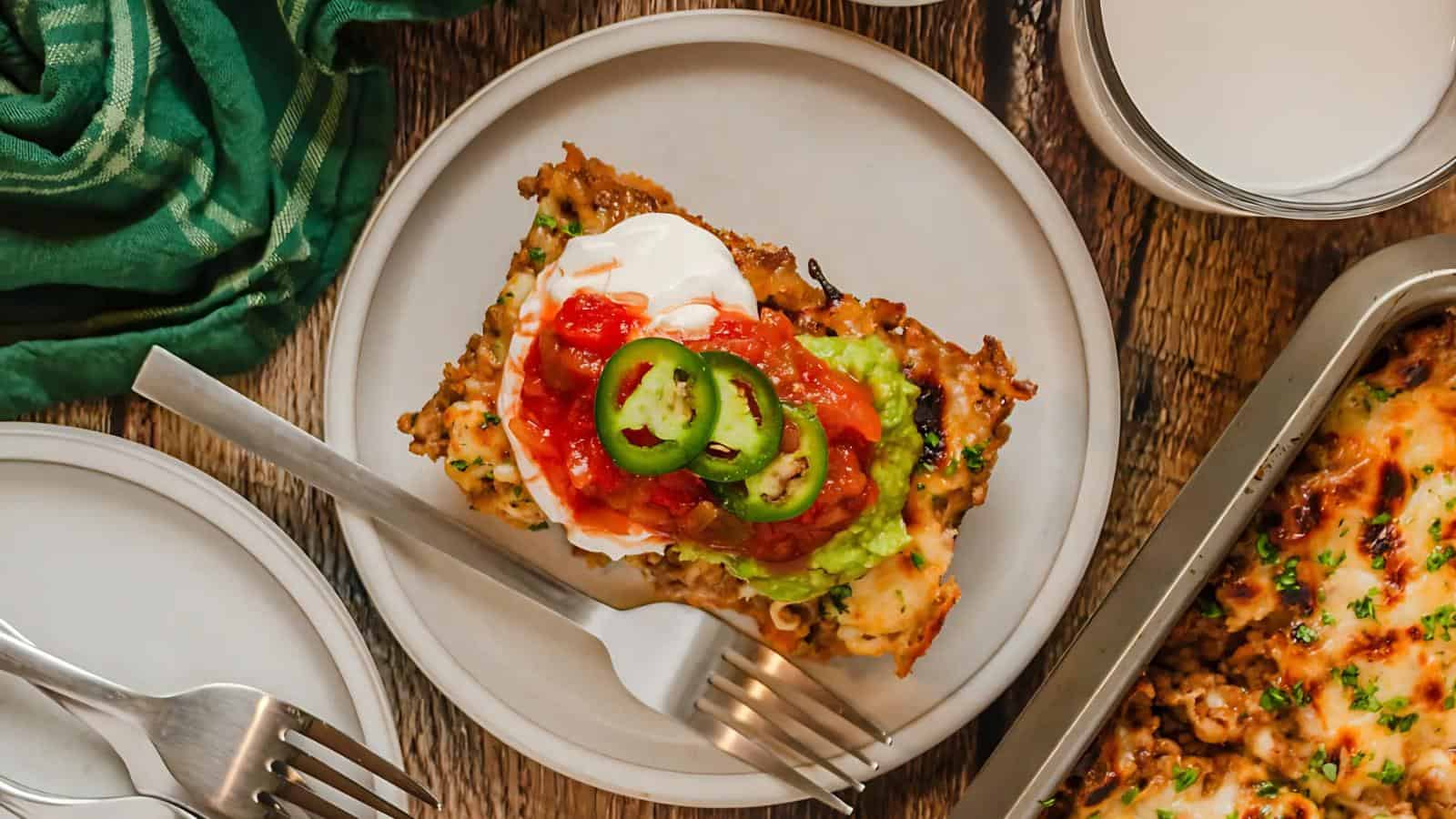 A plate with a serving of layered casserole topped with sour cream, guacamole, tomato salsa, and jalapeños. Forks and a green cloth are nearby.