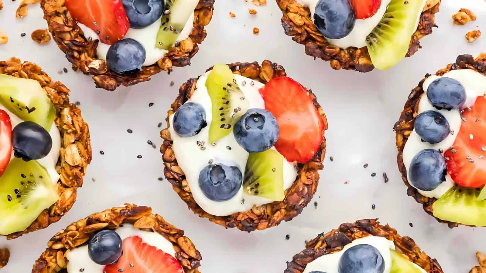 Granola tarts topped with yogurt, blueberries, sliced strawberries, and kiwi, arranged on a white surface.