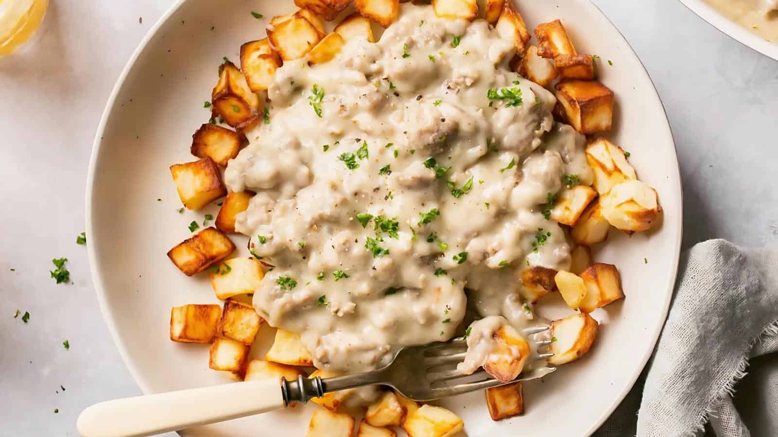 A plate of browned diced potatoes topped with creamy sausage gravy, garnished with chopped herbs. A fork rests on the side.