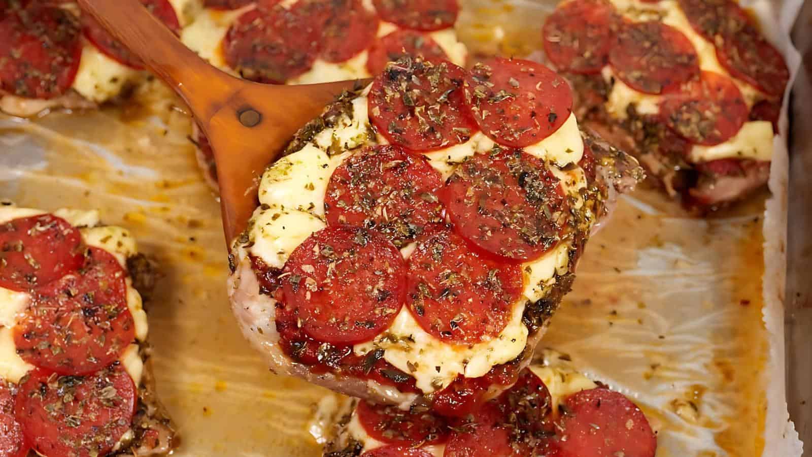 Close-up of a baked chicken dish topped with melted cheese, pepperoni slices, and sprinkled herbs, being served with a wooden spatula.