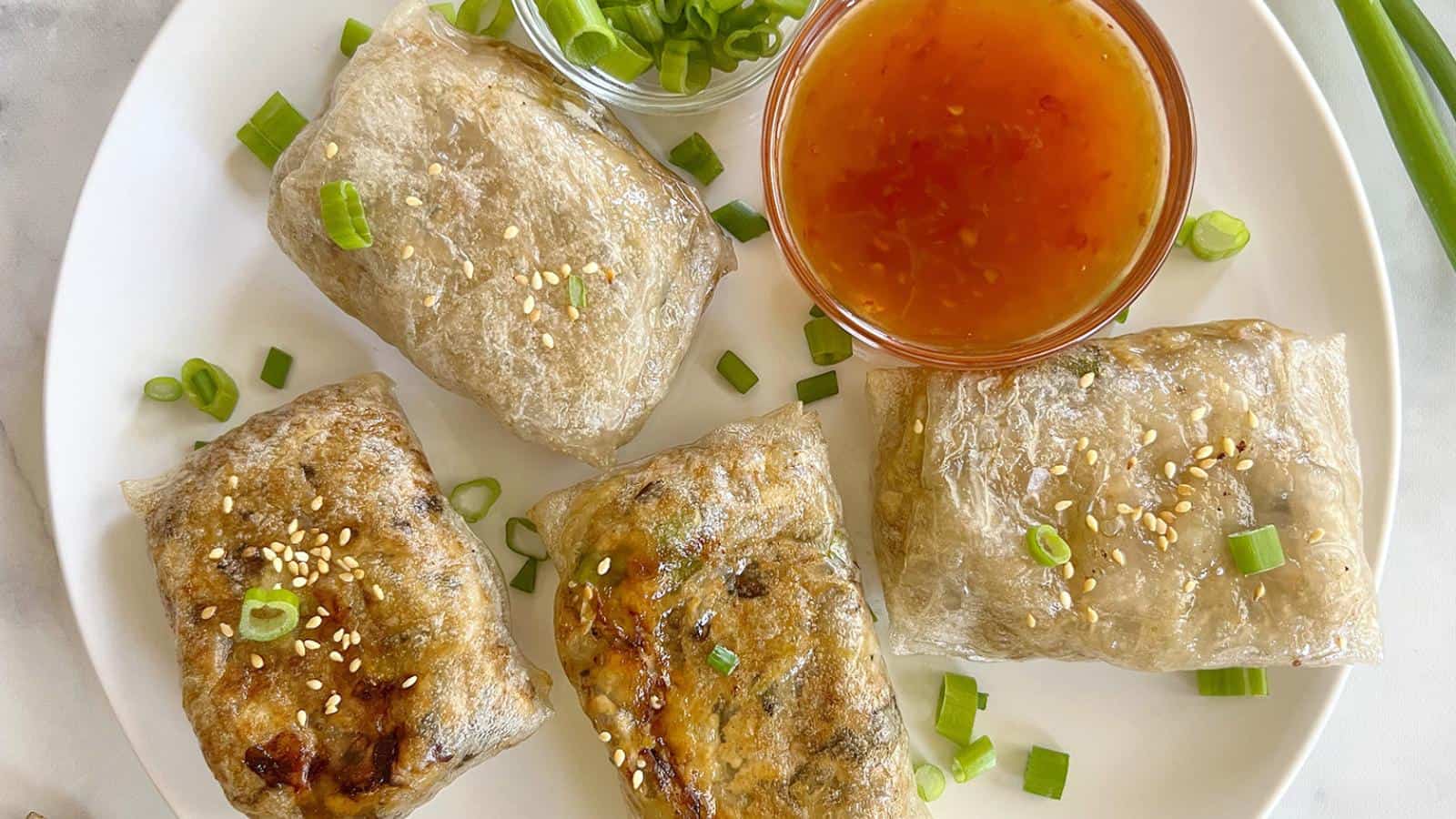 Four spring rolls on a plate, garnished with sesame seeds and green onions, served with a small bowl of dipping sauce.