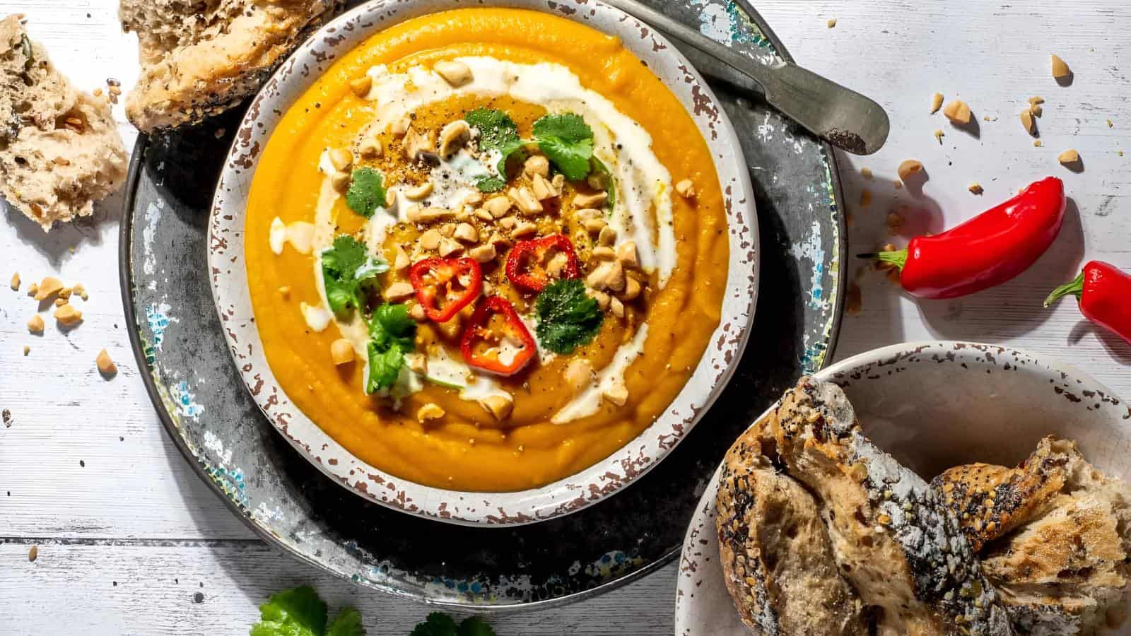 A bowl of creamy orange soup topped with sliced red chilies, nuts, and herbs, accompanied by a side of rustic bread and red peppers.