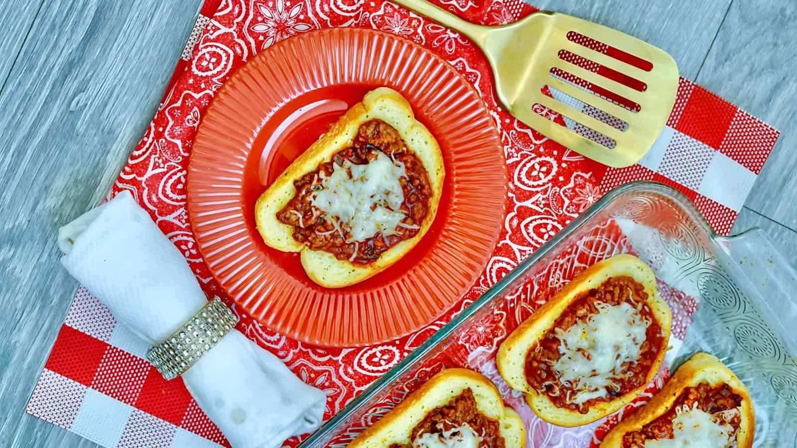 Slices of bread topped with tomato sauce and cheese on a red plate and a baking dish, placed on a red patterned cloth. A gold spatula and napkin are nearby.