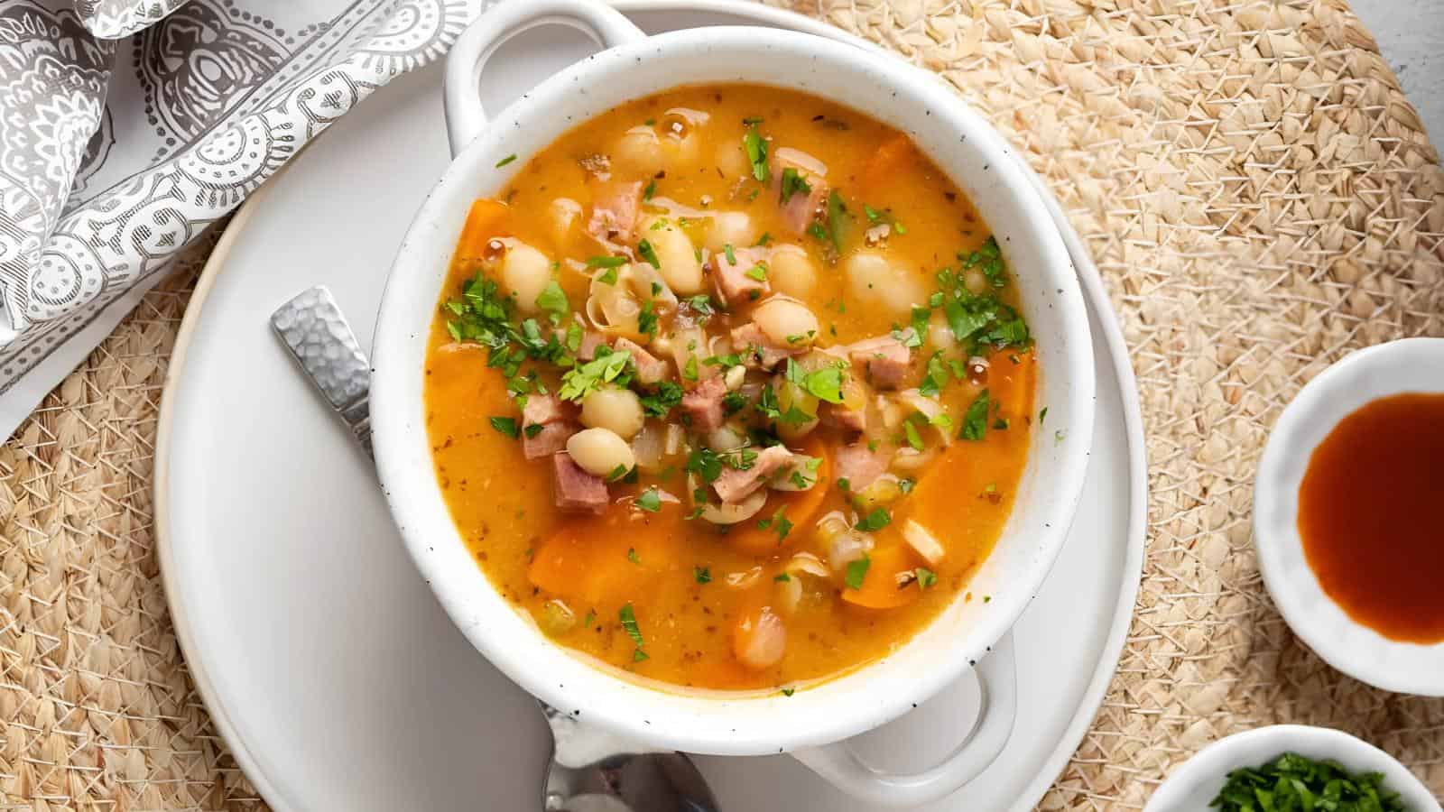 A white bowl of soup with white beans, diced meat, and herbs, garnished with parsley. It is placed on a plate with a spoon, sitting on a woven surface with a small dish of sauce nearby.