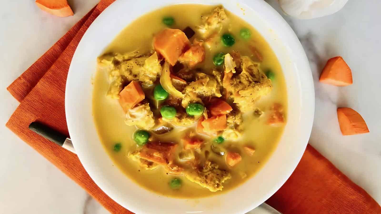 A bowl of yellow curry with chicken, sweet potatoes, and peas on a white plate, placed on an orange napkin, with pieces of sweet potato scattered around.