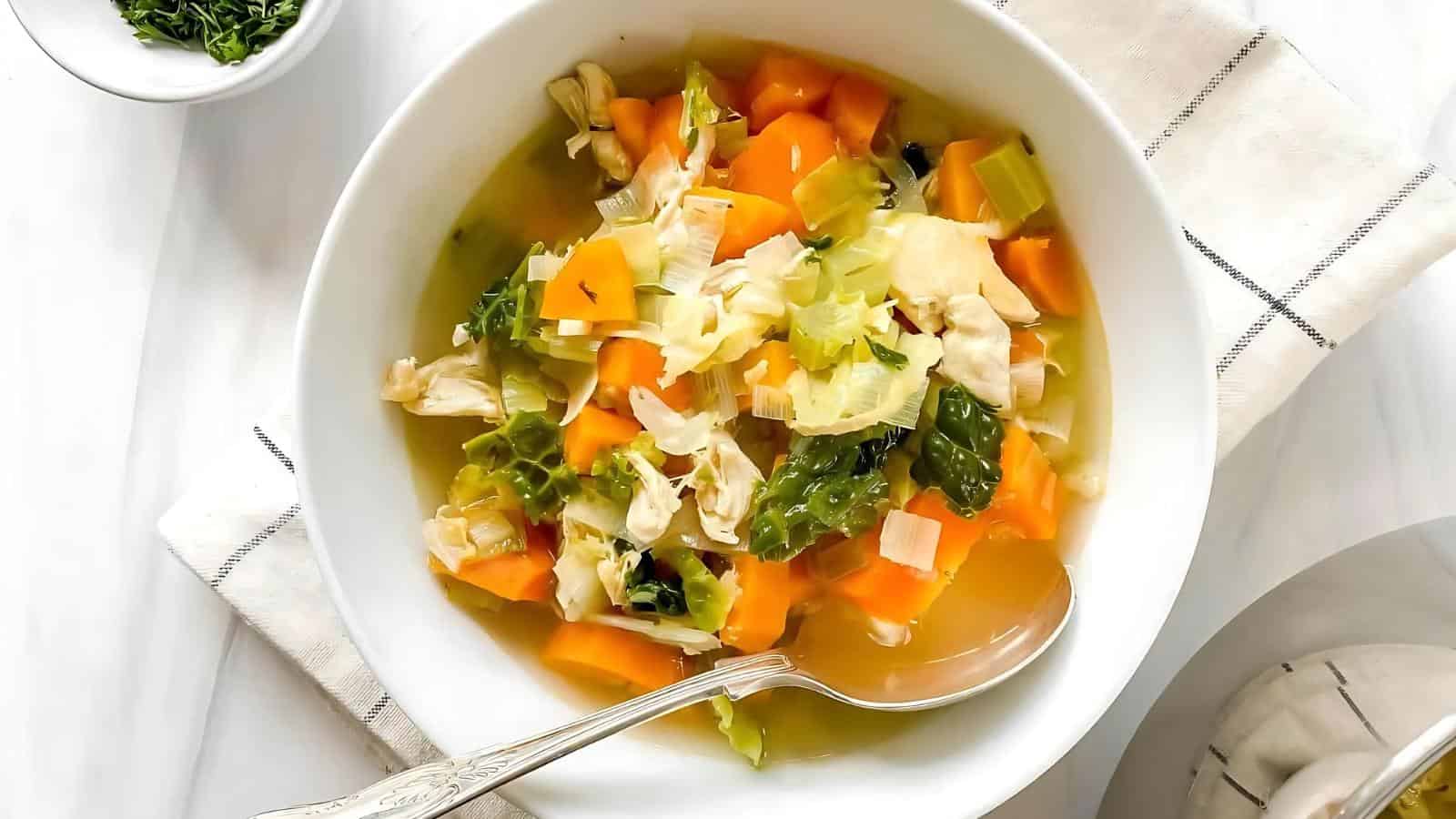 A bowl of vegetable soup with carrots, cabbage, celery, and greens, placed on a white surface with a spoon and napkin beside it.