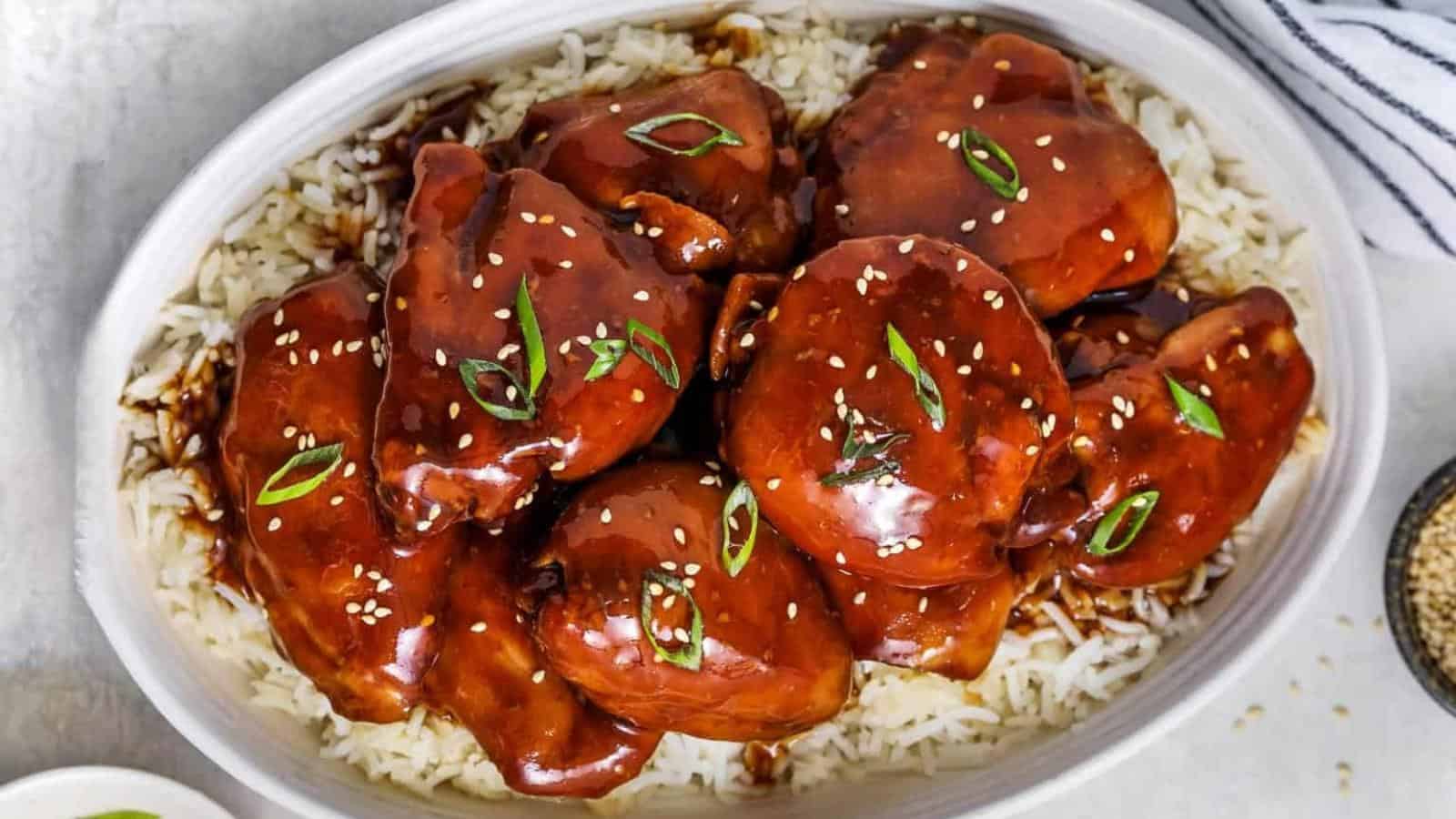 Chicken thighs glazed with a dark sauce, garnished with sesame seeds and green onions, served on a bed of white rice in an oval dish.