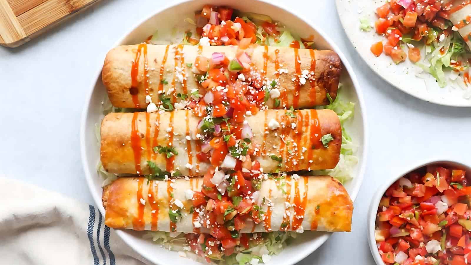 A plate of four crispy taquitos topped with diced tomatoes, onions, cheese, and drizzled with hot sauce, served on a bed of shredded lettuce.