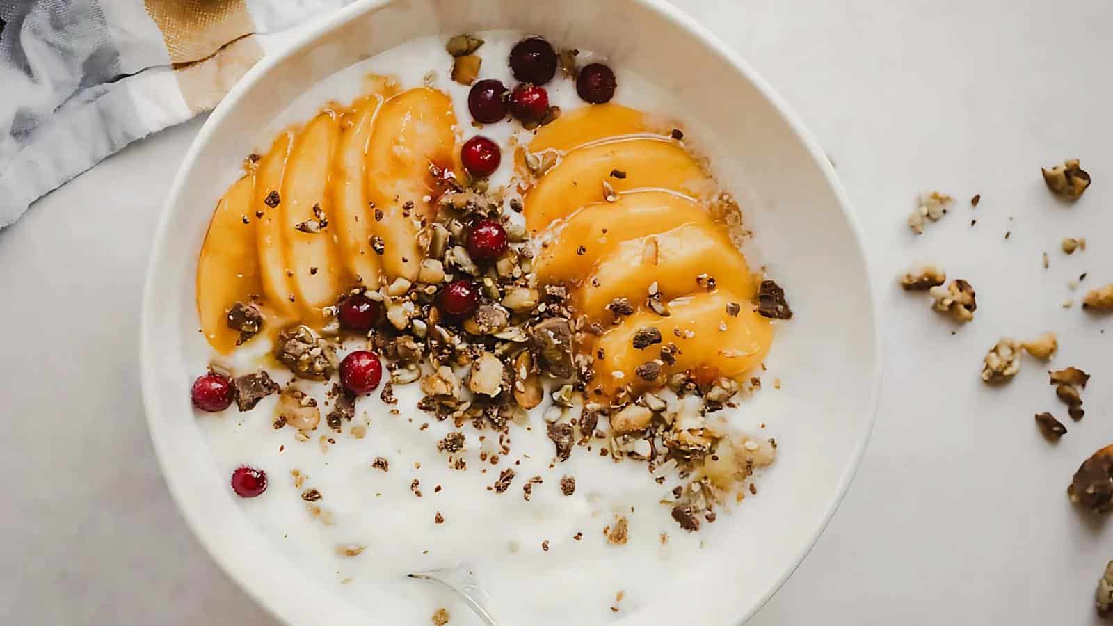 A bowl of yogurt topped with sliced peaches, cranberries, crushed nuts, and granola on a light surface.