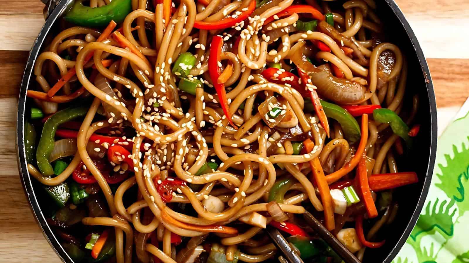 Stir-fried noodles with bell peppers, carrots, and mushrooms, topped with sesame seeds in a black pan.