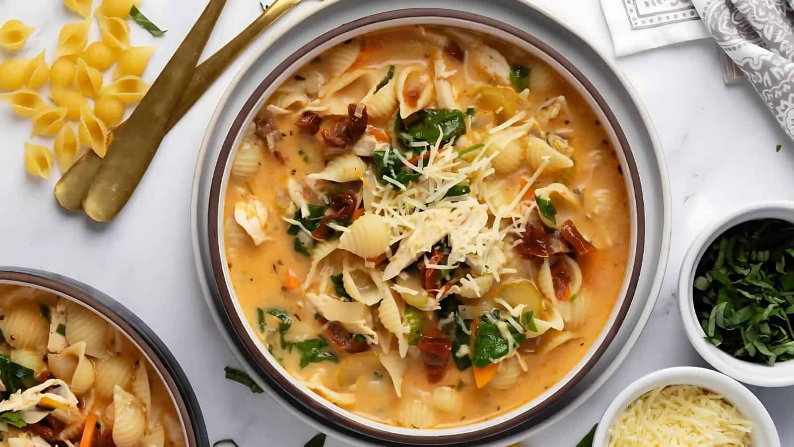 A bowl of creamy chicken soup with pasta shells, spinach, and shredded cheese. Garnished with sun-dried tomatoes. Nearby are uncooked pasta, herbs, and utensils on a white surface.