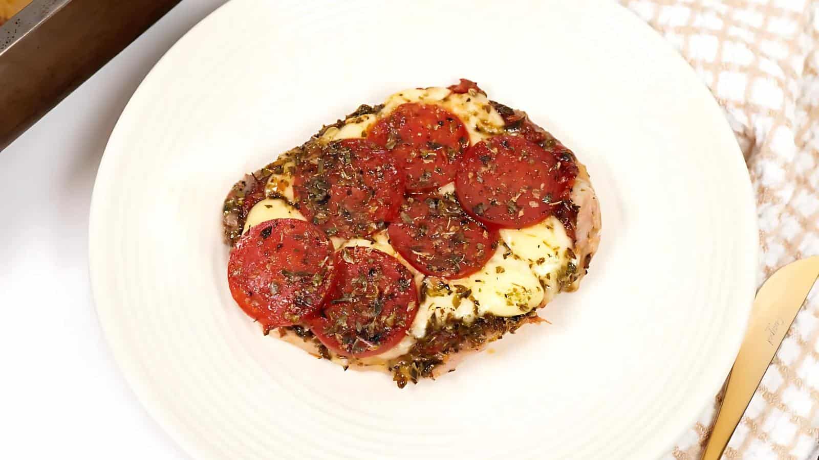 A square slice of pizza on a white plate, topped with pepperoni slices, melted cheese, and herbs.