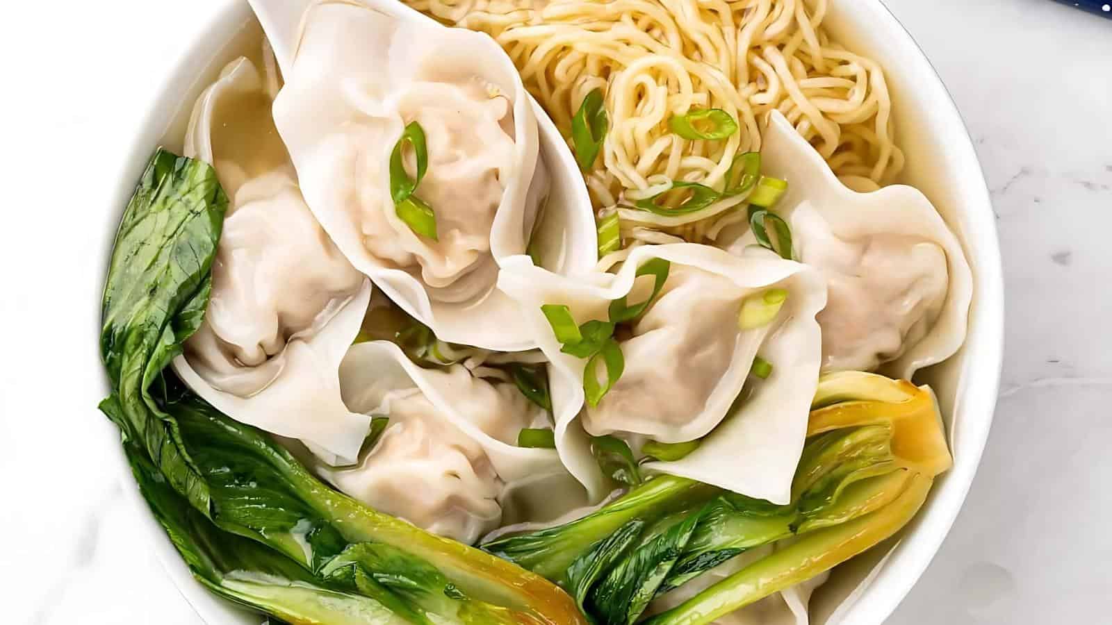 A bowl of noodle soup with dumplings, bok choy, and sliced spring onions.