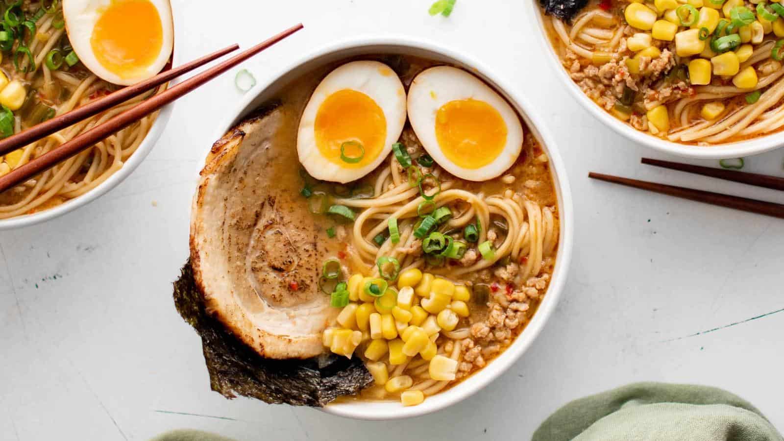 Bowl of ramen with sliced pork, boiled egg halves, corn, green onions, and seaweed. Chopsticks rest on the bowl's edge. Another bowl is partially visible in the background.