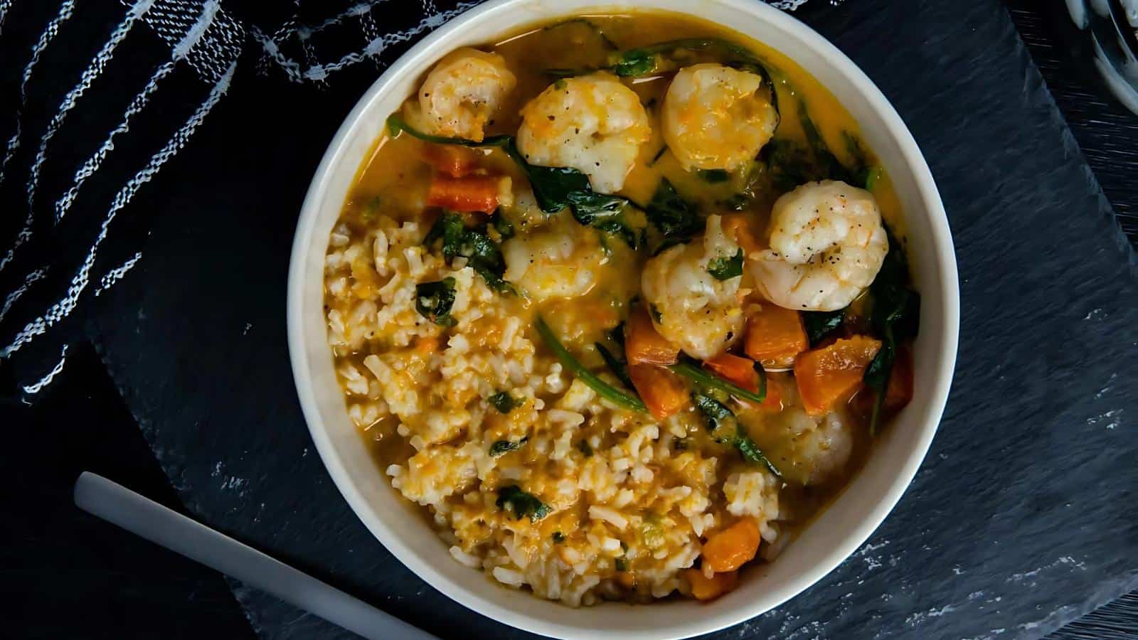 Bowl of shrimp curry with rice, spinach, and diced carrots on a black slate surface, accompanied by a striped cloth napkin.