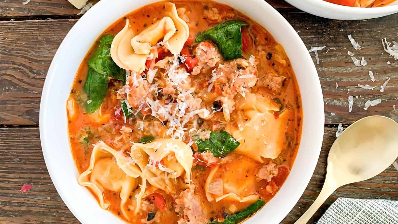A bowl of creamy tomato tortellini soup with spinach, sausage, and grated cheese, placed on a wooden table beside a spoon.
