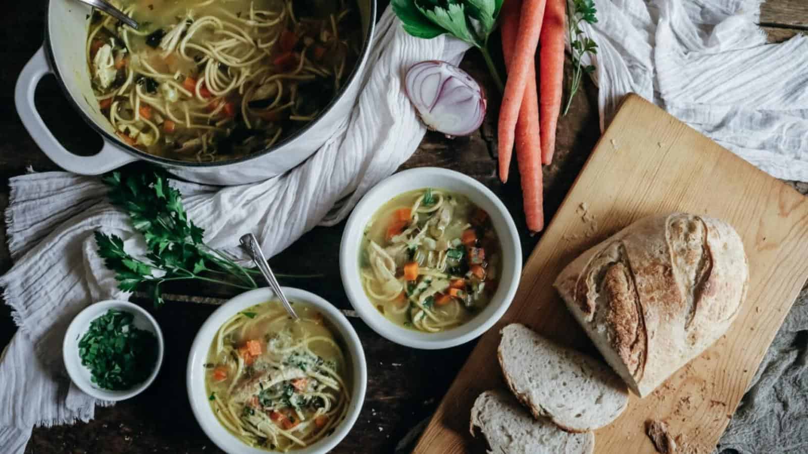 A pot of noodle soup with vegetables, two bowls of soup, a loaf of bread, sliced pieces, carrots, onion, and parsley on a wooden table.